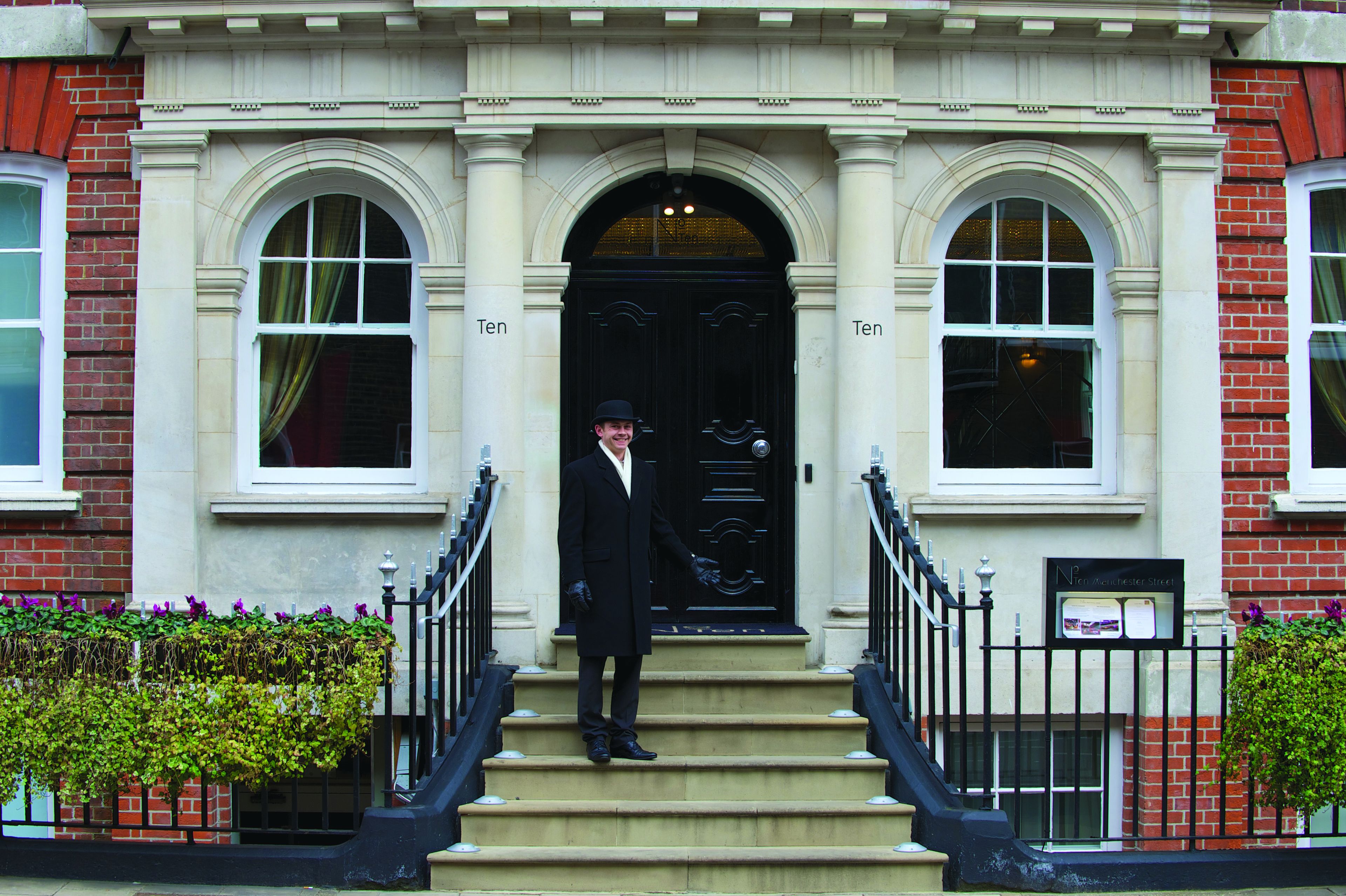 A man standing on the steps of a building with a black door and two arched windows on either side.