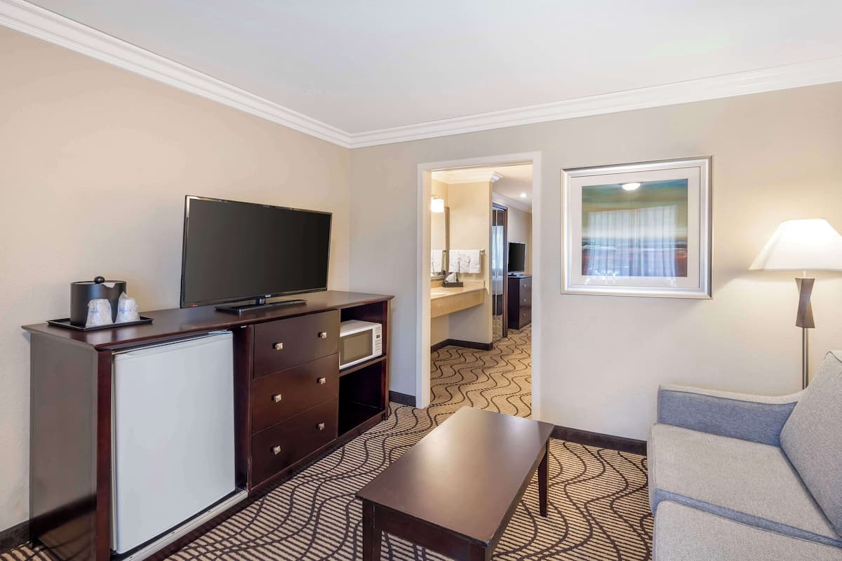 A hotel room with a flat-screen TV on a dark wooden cabinet, a small refrigerator, a sofa, a table, and a framed picture on the wall.