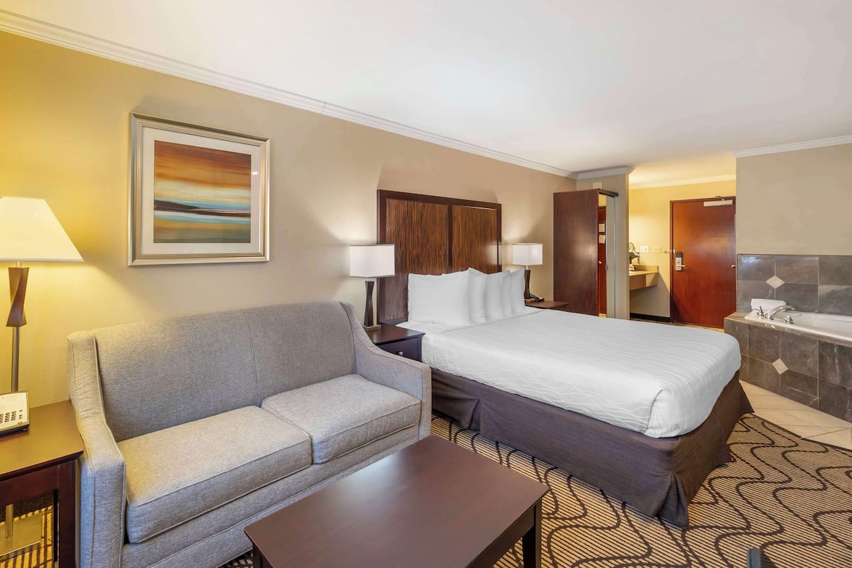 A hotel room with a bed, a sofa, a desk, a lamp, a painting, and a bathroom with a bathtub and sink.