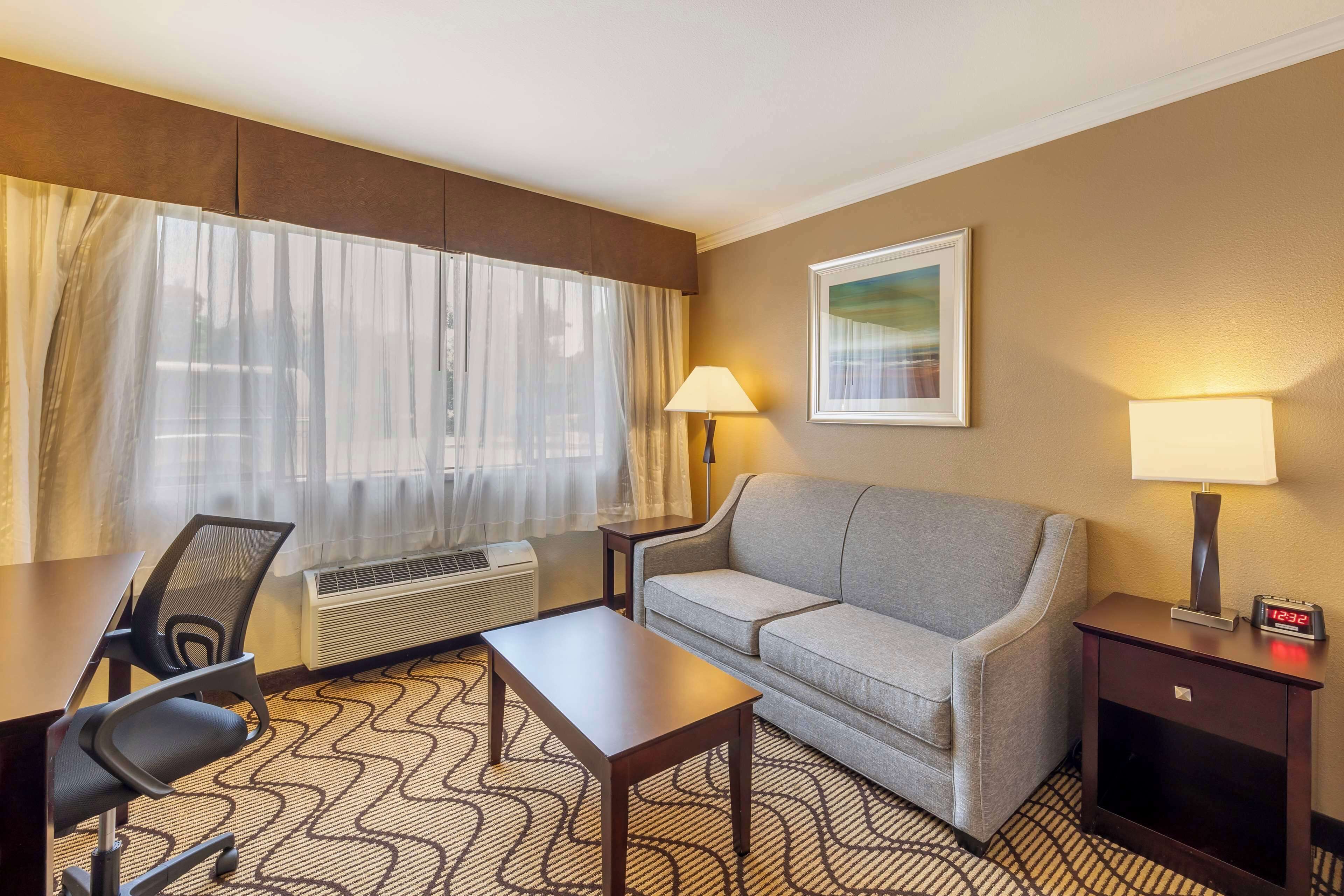 A hotel room with a sofa, a desk, a chair, two lamps, a clock, and a framed picture on the wall.