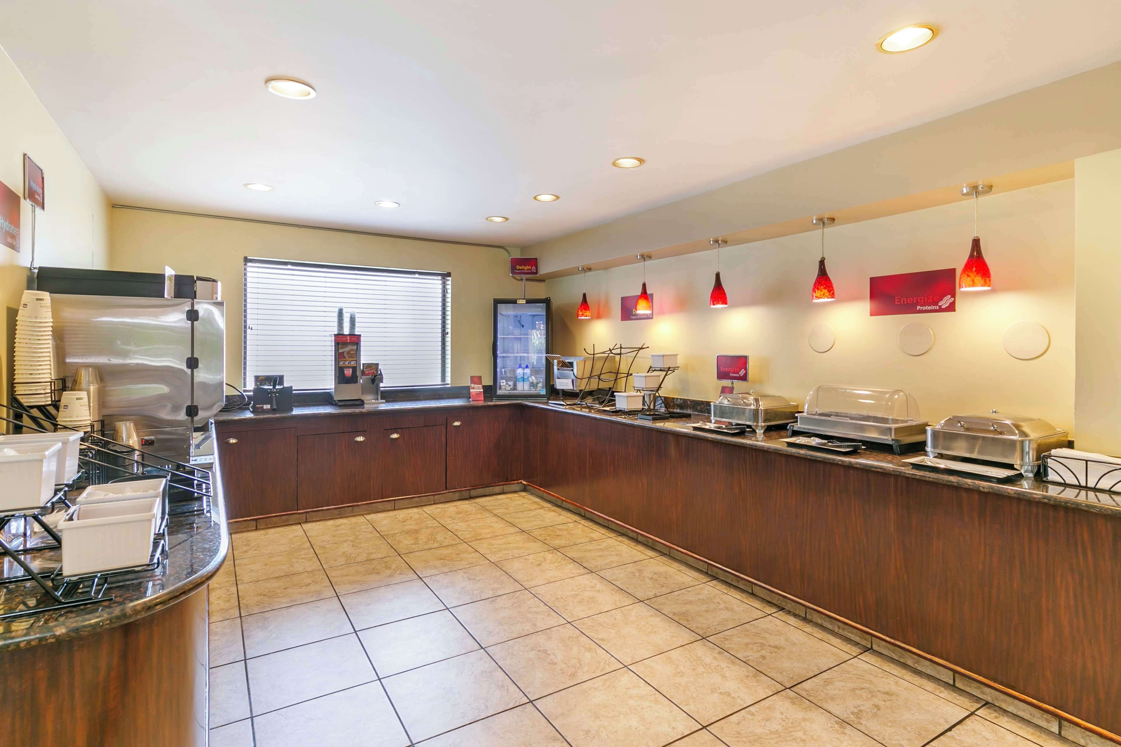 Zona de desayuno de hotel con mostrador, cafeteras y un refrigerador.