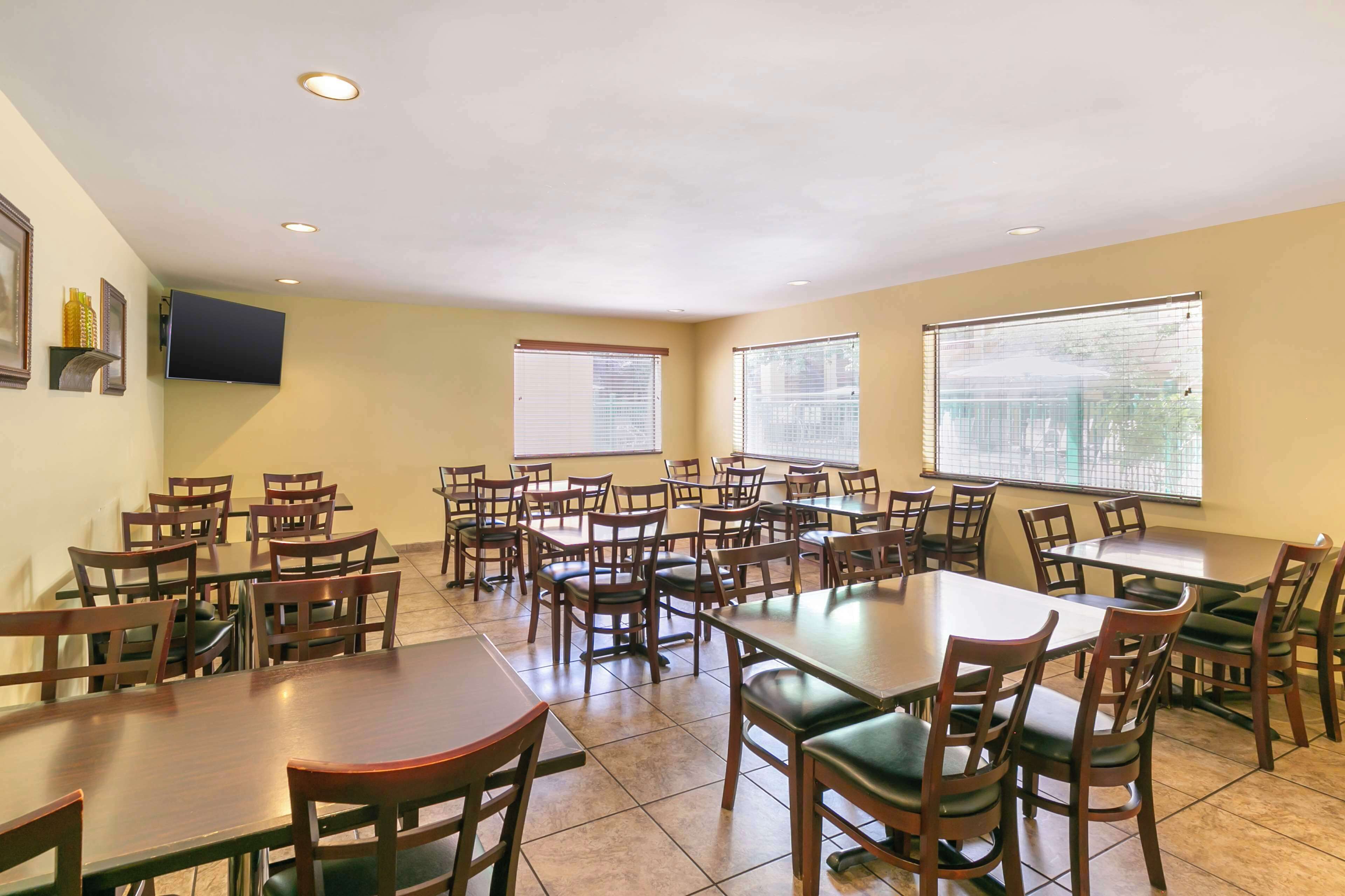 A dining area with multiple tables and chairs, a flat-screen TV mounted on the wall, and large windows with blinds.
