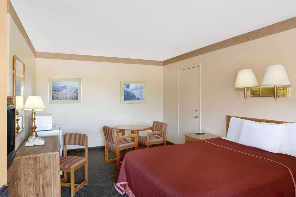 Habitación de hotel con una cama grande, un escritorio con lámpara, dos sillas, una mesita y dos lámparas de pared.