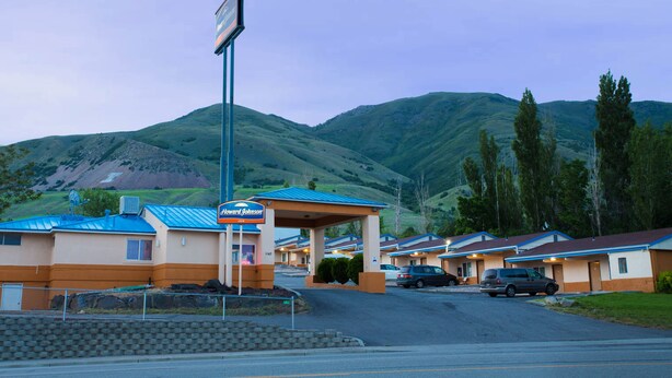 A motel with a blue roof and beige walls, a covered parking area, and a mountainous backdrop. - Howard Johnson by Wyndham Brigham City (Brigham City)