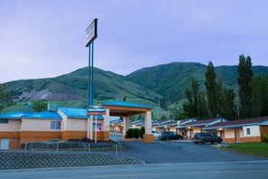 Ein Motel mit blauem Dach und beige farbenen Wänden, einem überdachten Parkplatz und einer Bergkulisse im Hintergrund.