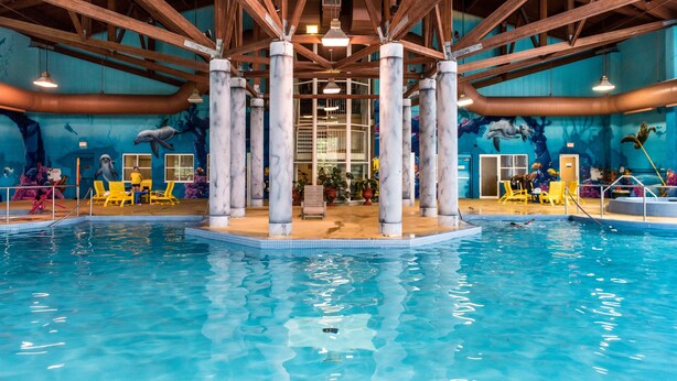 An indoor swimming pool with clear blue water, wooden beams, and yellow lounge chairs. - Deerhurst Resort (Huntsville)