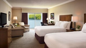 A hotel room with two beds, a desk, a chair, a TV, and a balcony with a view of greenery. - Deerhurst Resort (Huntsville)