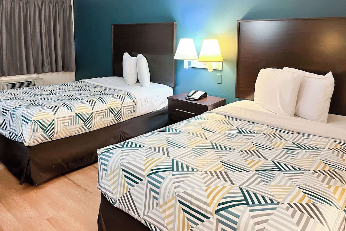 A hotel room with two beds, a nightstand with a phone, and a wall-mounted lamp.