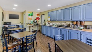 Una cocina con gabinetes azules, mesas y sillas de madera. Hay un microondas, un tostador y diversos electrodomésticos de cocina sobre la encimera.