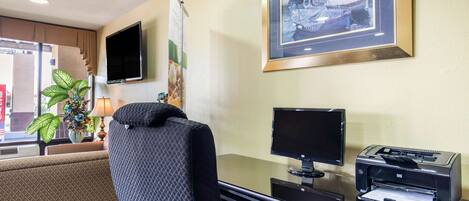 A hotel room with a desk, a computer, a printer, a TV, and a framed picture on the wall.