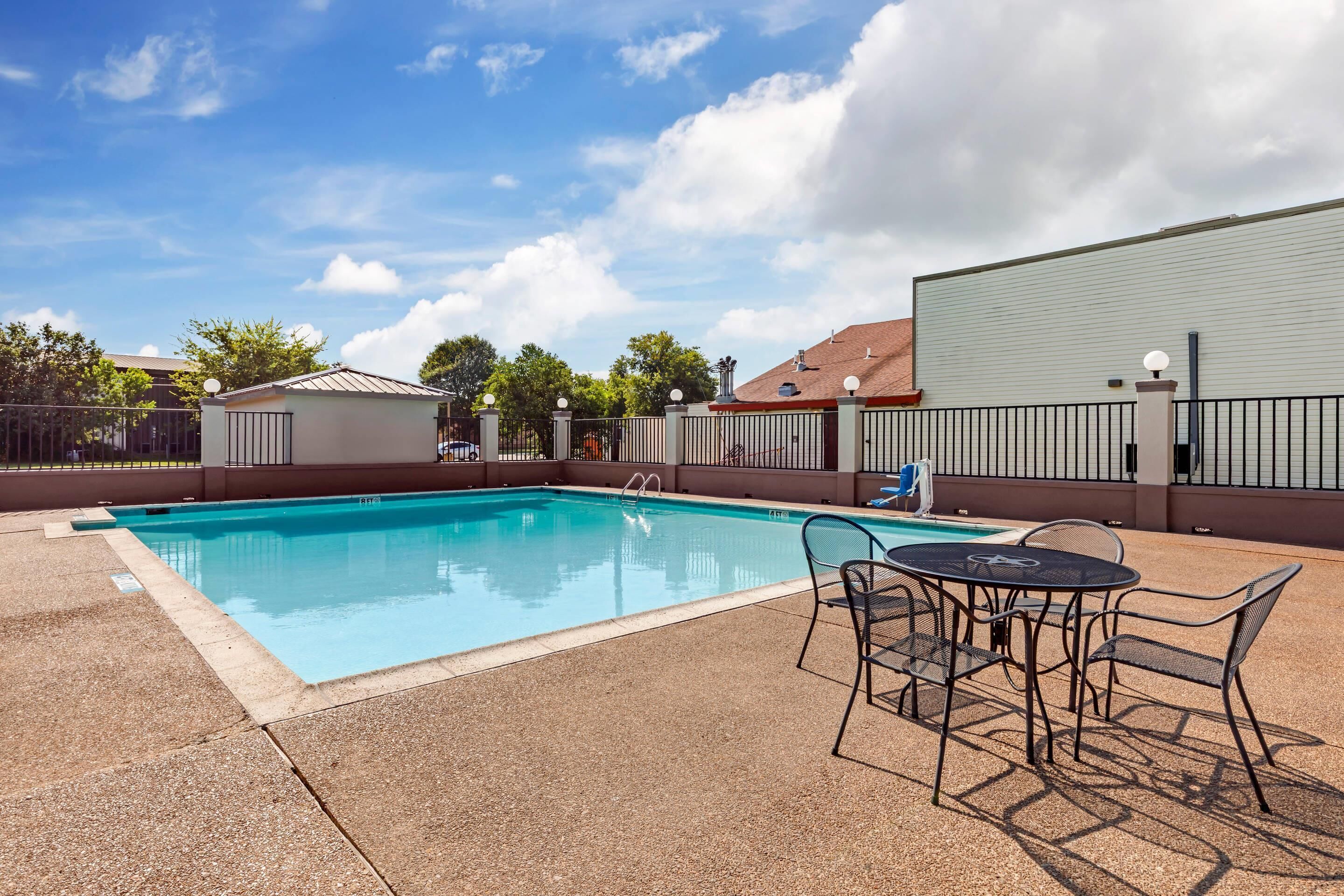A rectangular swimming pool with a tiled deck, surrounded by a metal fence. There is a table with chairs and a blue lounge chair near the pool.