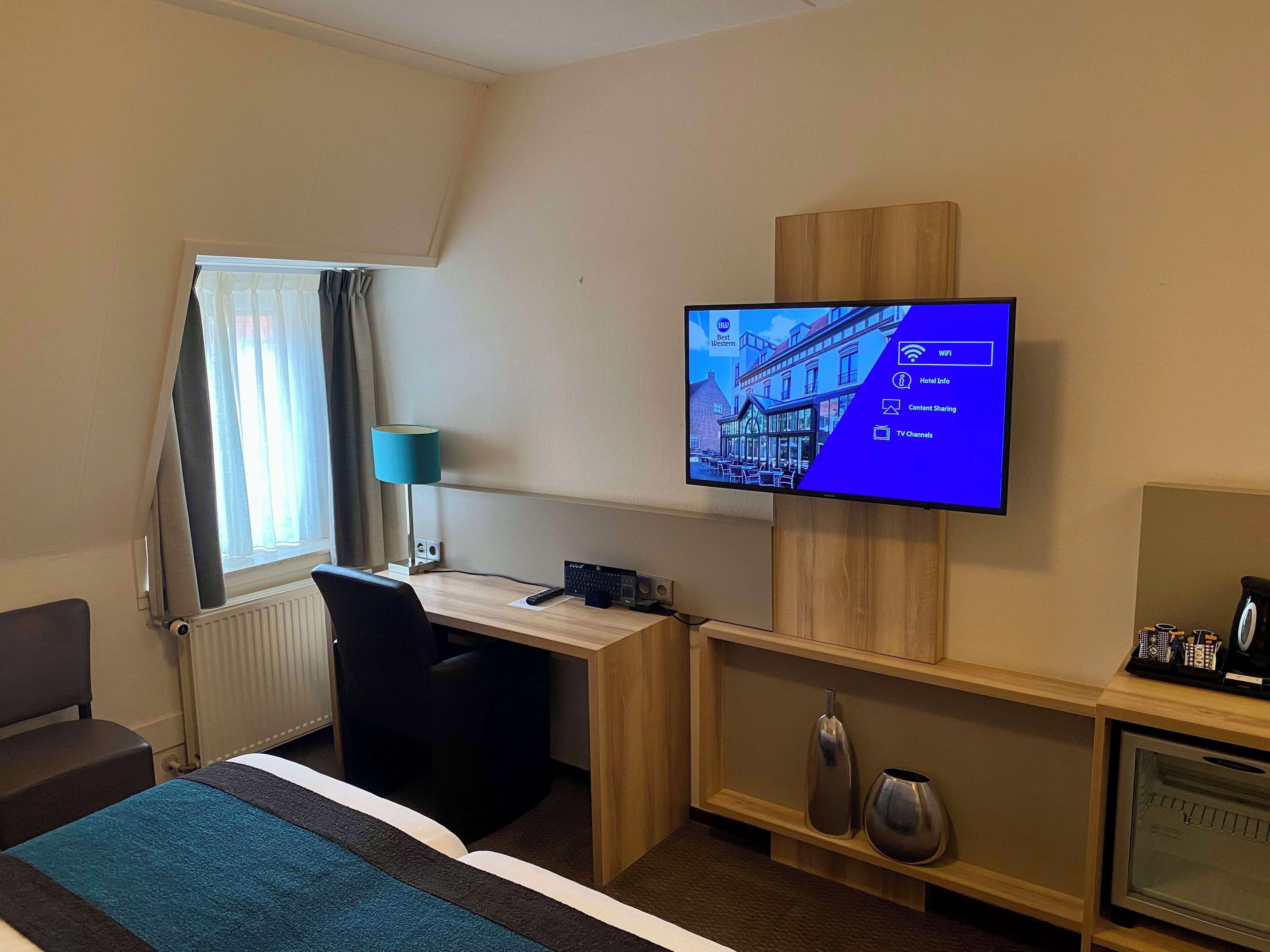 A hotel room with a bed, a desk, a television mounted on the wall, a lamp, and a small cabinet with a coffee maker.