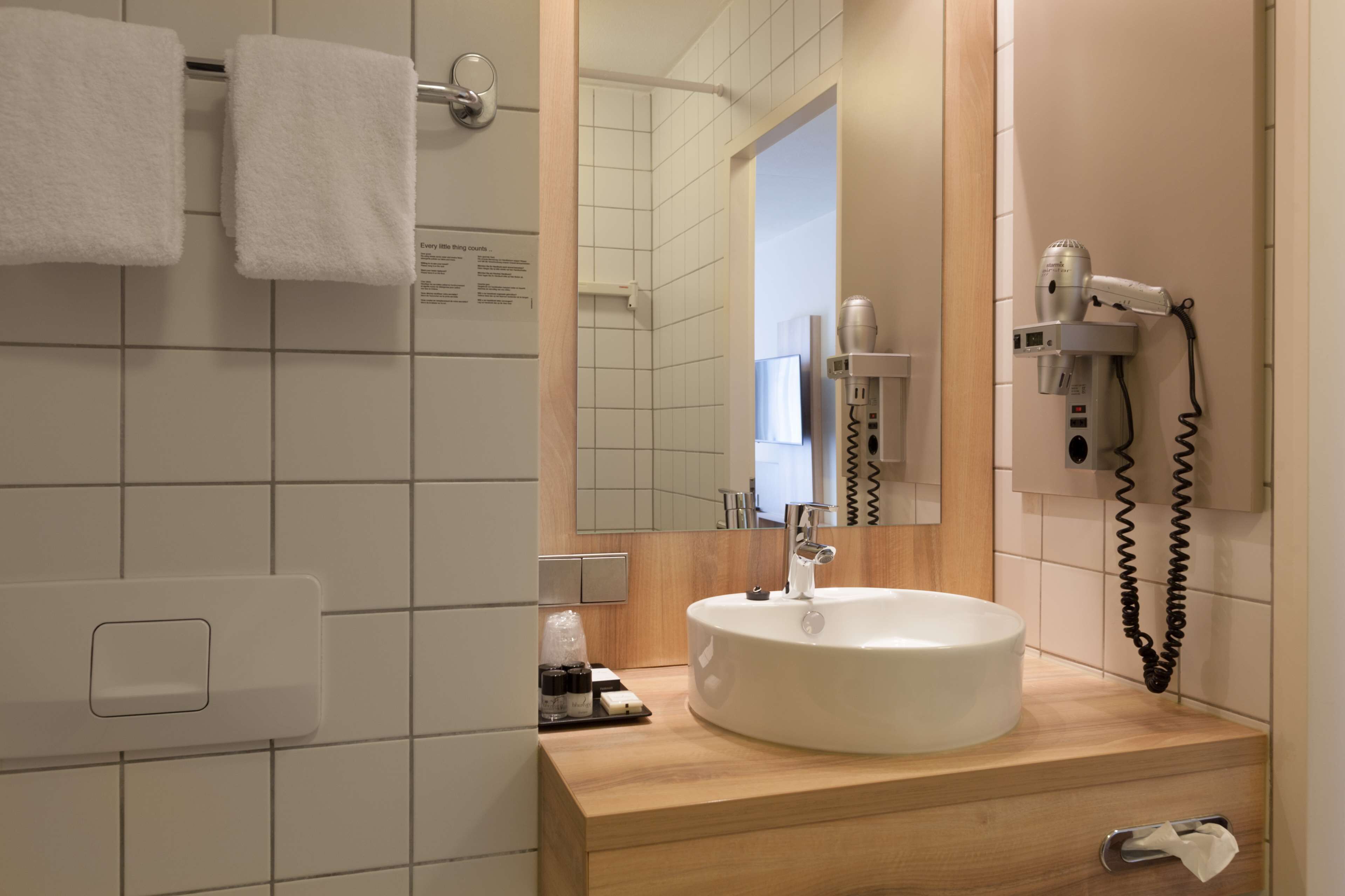Un baño moderno con un lavamanos blanco, un espejo, un secador de pelo y un tendedero para toallas.