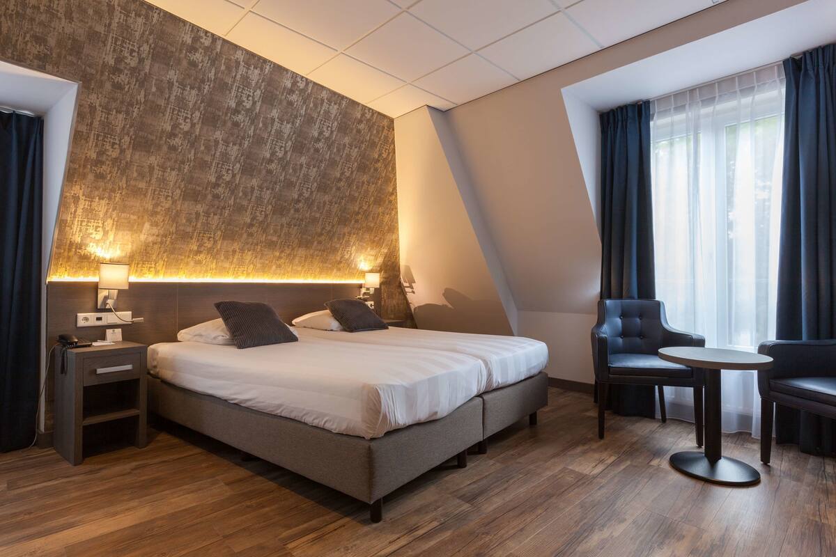 Habitación de hotel moderna con una cama grande, una mesita pequeña, dos sillas, una mesita de noche y una pared grande con textura.