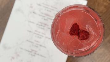 Un vaso con una bebida rosada y dos fresas sobre hielo, colocado sobre una mesa de madera con un menú al fondo.