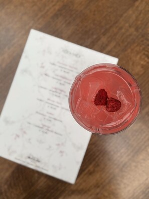 Un verre contenant une boisson rose et deux fraises sur de la glace, posé sur une table en bois, avec un menu en arrière-plan.