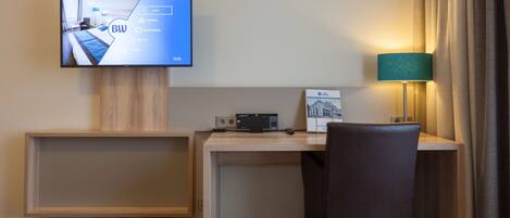 A hotel room with a TV displaying hotel information, a desk with a keyboard and mouse, a chair, and a lamp.