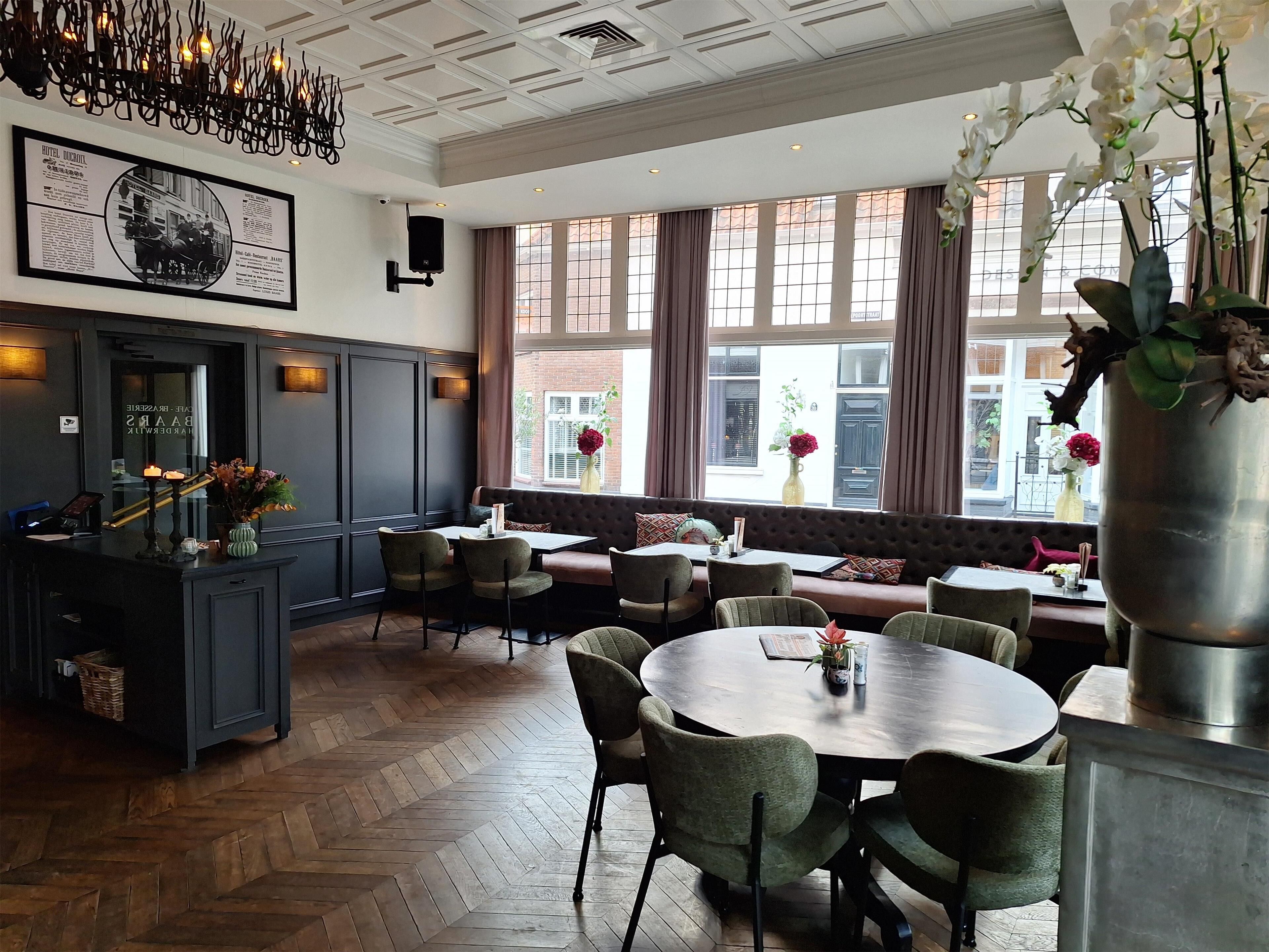 A restaurant with a herringbone wood floor, a bar area with a mirror, and a large window with curtains.