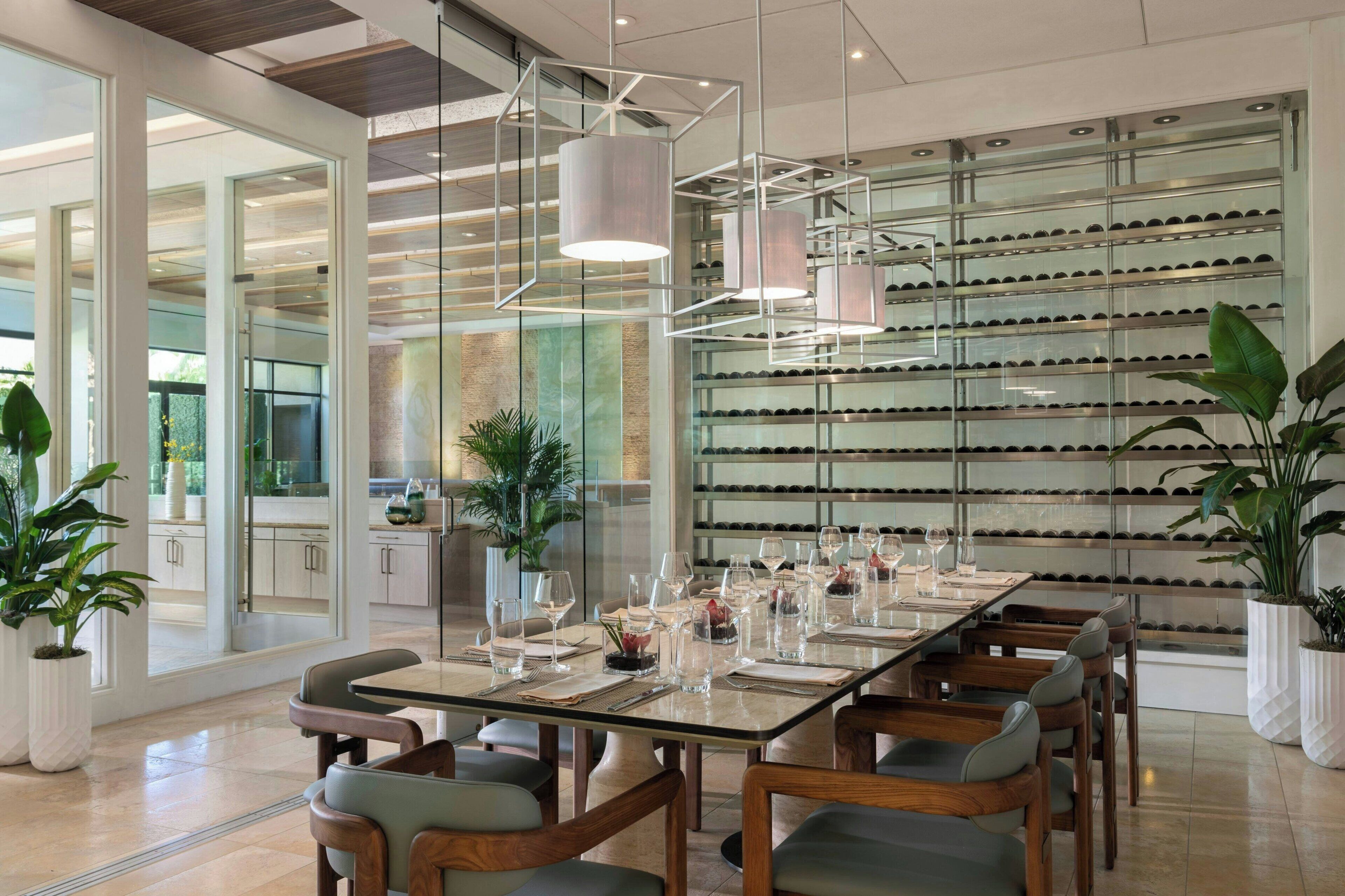 Un espace de salle à manger moderne, avec une longue table dressée pour un repas, entourée de chaises. On aperçoit, à l’arrière-plan, des plantes en pot et un meuble à vin.
