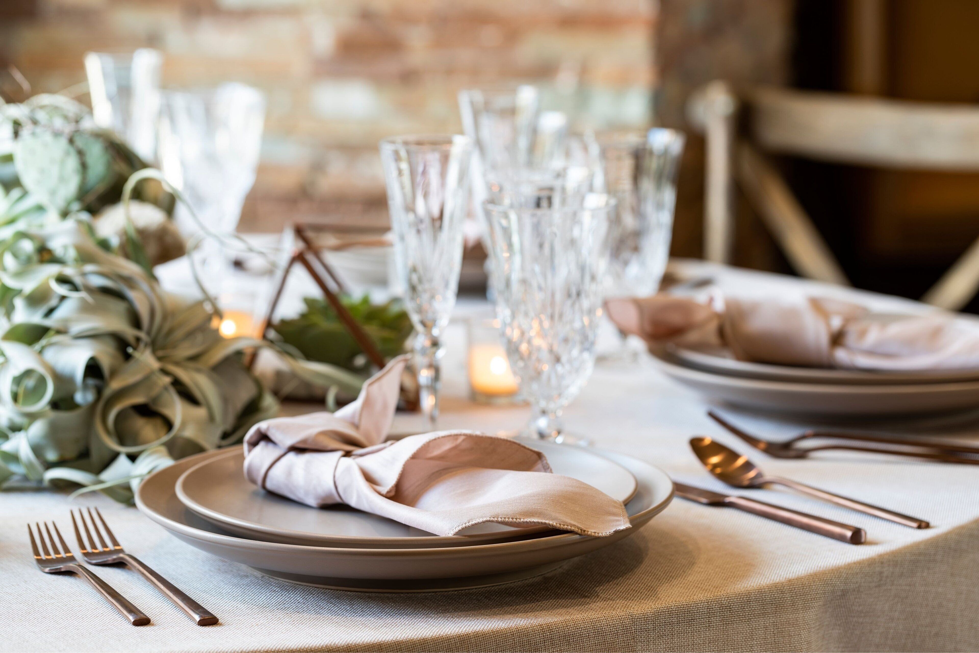 A table set with plates, cutlery, and glasses, with a candle and greenery in the background.