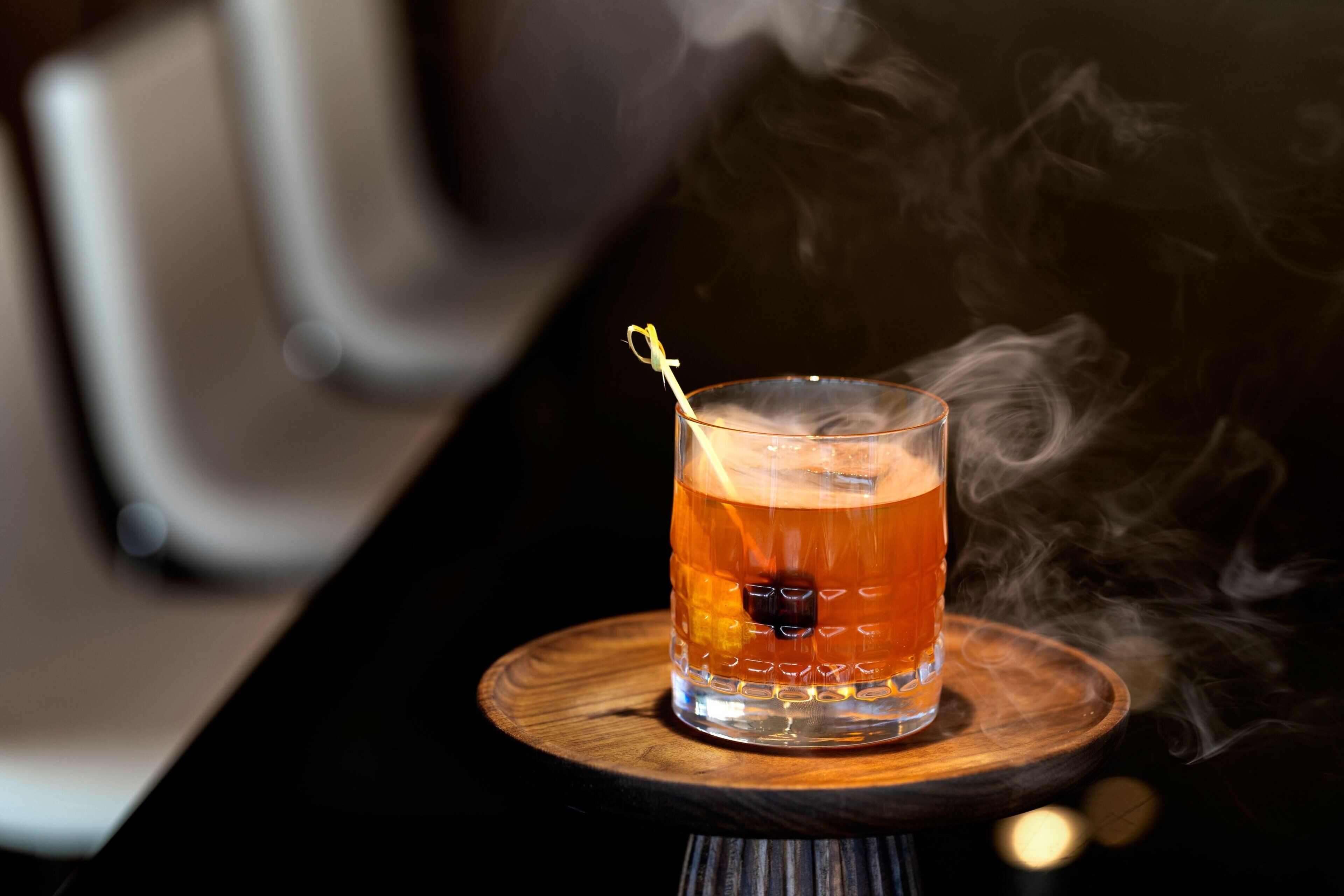 Un verre contenant un liquide ambré, avec une paille, posé sur un dessous de verre en bois. De la vapeur s’en échappe et s’élève au-dessus du verre, le tout étant placé sur une table.