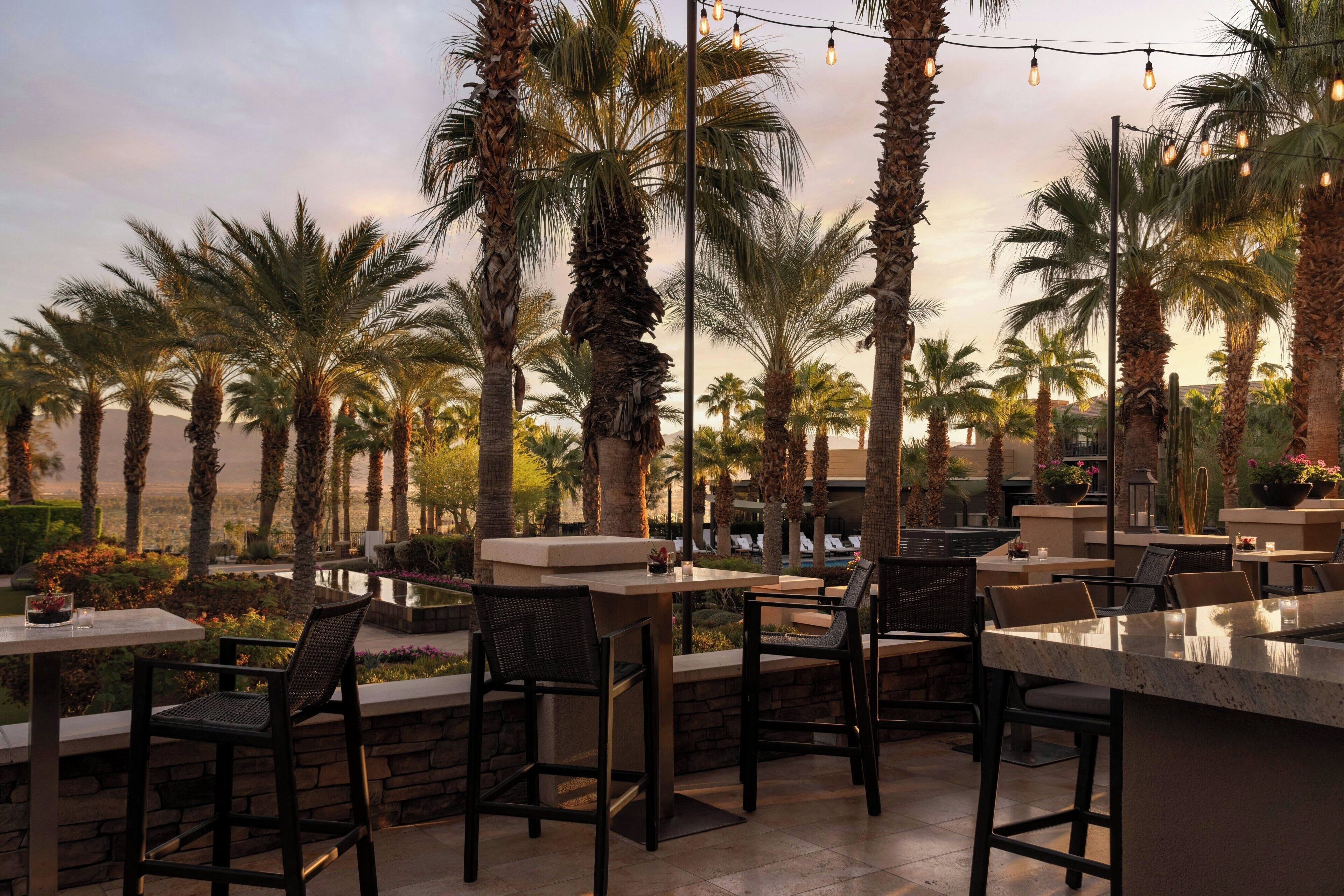 An outdoor patio area with high tables and chairs, surrounded by palm trees and a view of a pool.