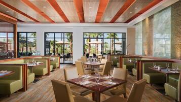 A spacious restaurant with high ceilings and large windows, offering views of palm trees and a clear sky.
