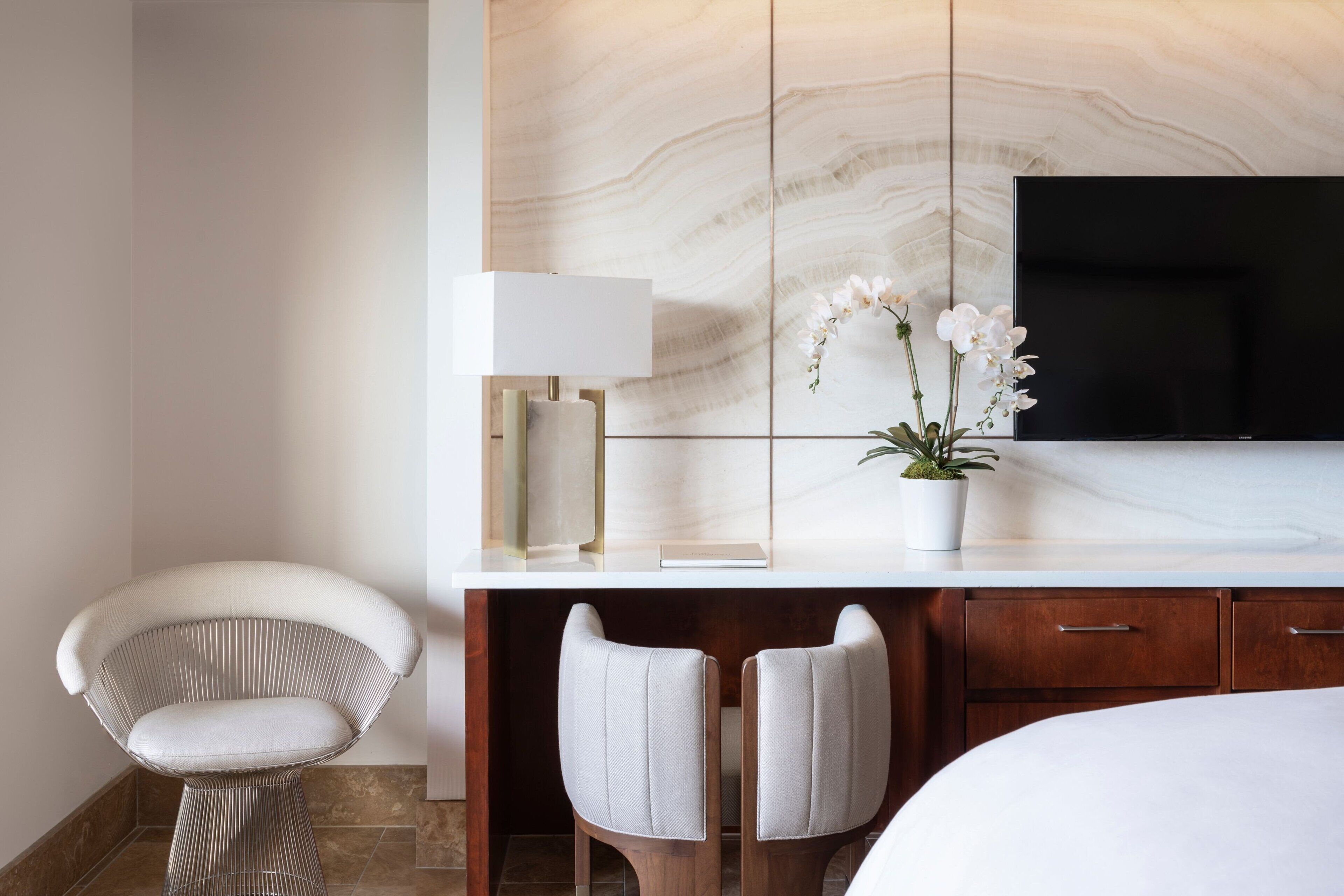 Habitación de hotel moderna con pared de mármol, televisor de pantalla plana, una silla blanca, dos taburetes altos y una mesita de noche con lámpara y planta de orquídea.