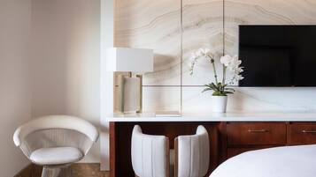 Habitación de hotel moderna con pared de mármol, televisor de pantalla plana, una silla blanca, dos taburetes altos y una mesita de noche con lámpara y planta de orquídea.