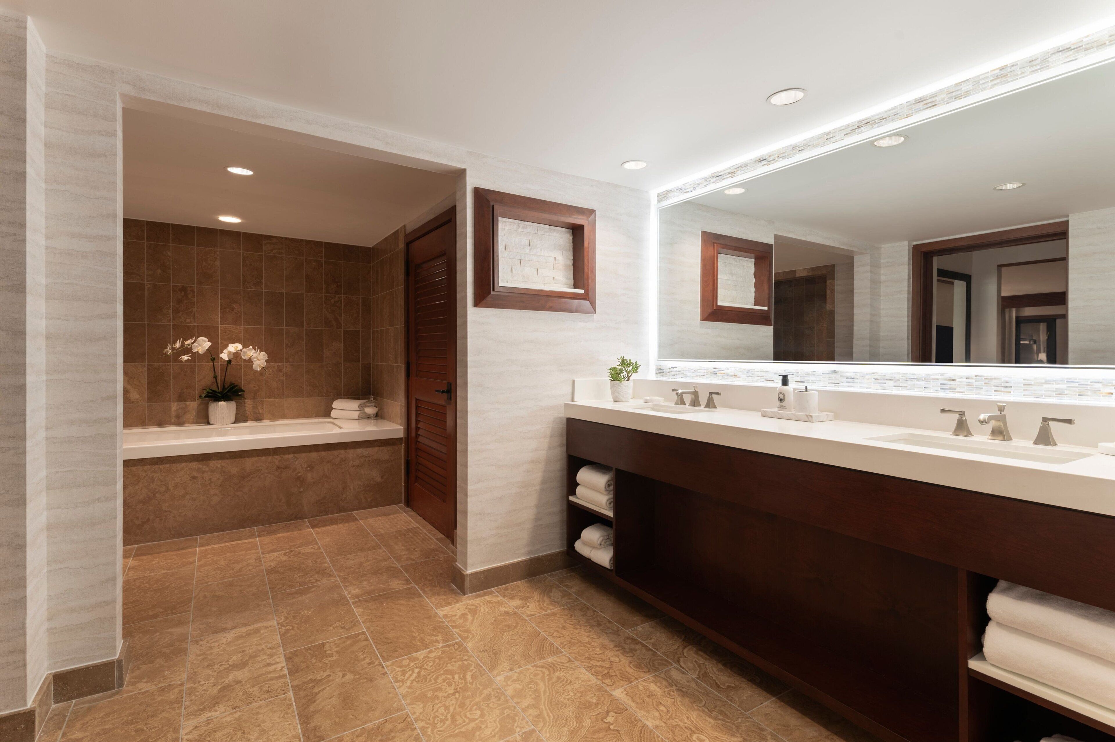 A modern bathroom with a large mirror, dual sinks, a bathtub, and a vanity with shelves.