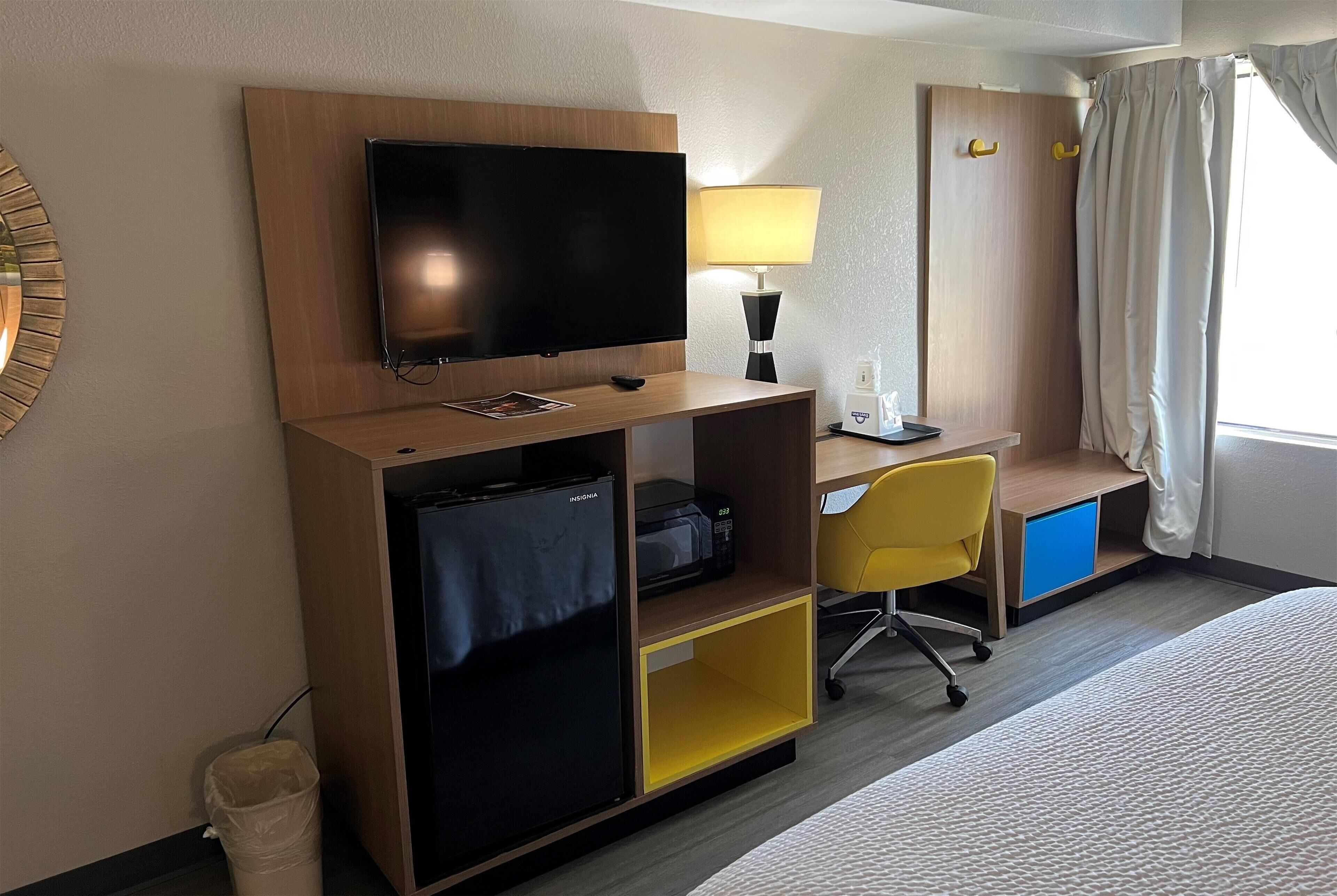 A hotel room with a flat-screen TV, a desk with a chair, a bed, a trash bin, and a window with curtains.