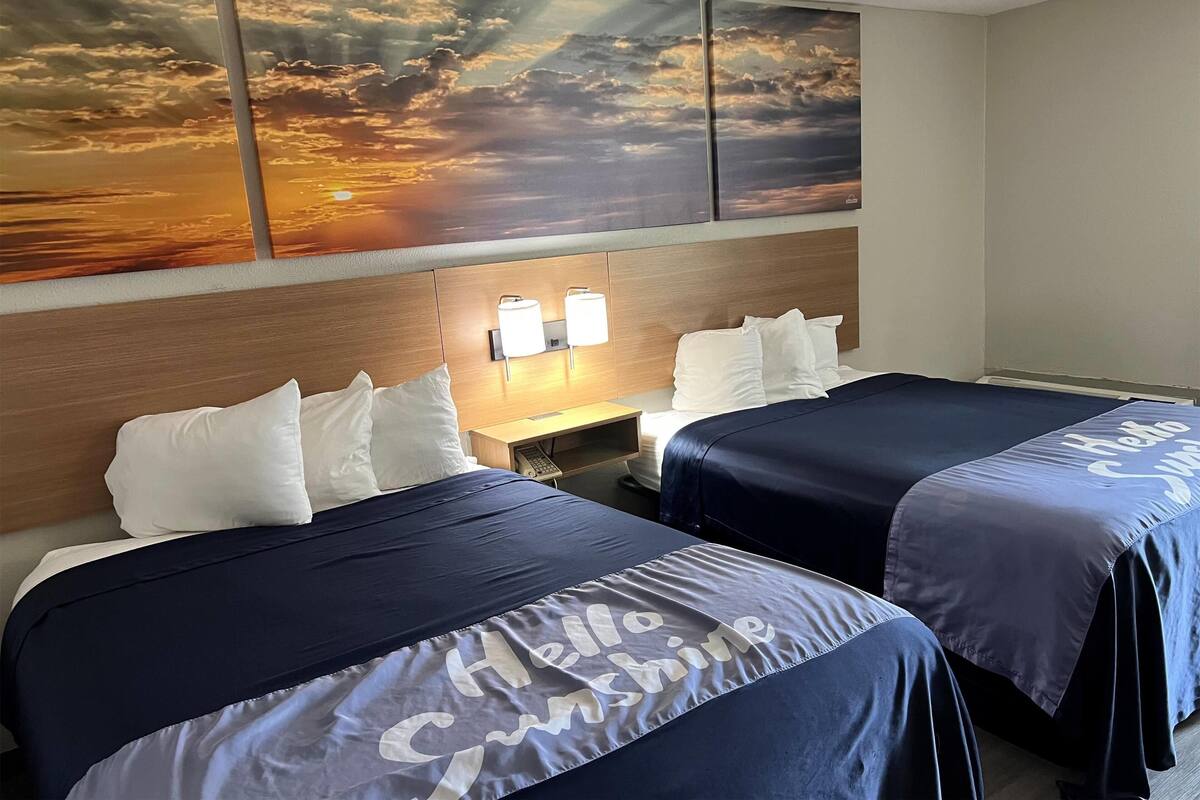 A hotel room with two beds, each with a 'Hello Sunshine' bedspread, a wooden headboard, a nightstand with a lamp, and a sunset mural on the wall.
