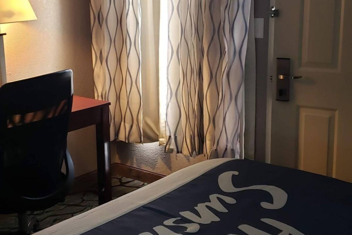 A hotel room with a bed, a desk, a chair, a lamp, and a door.