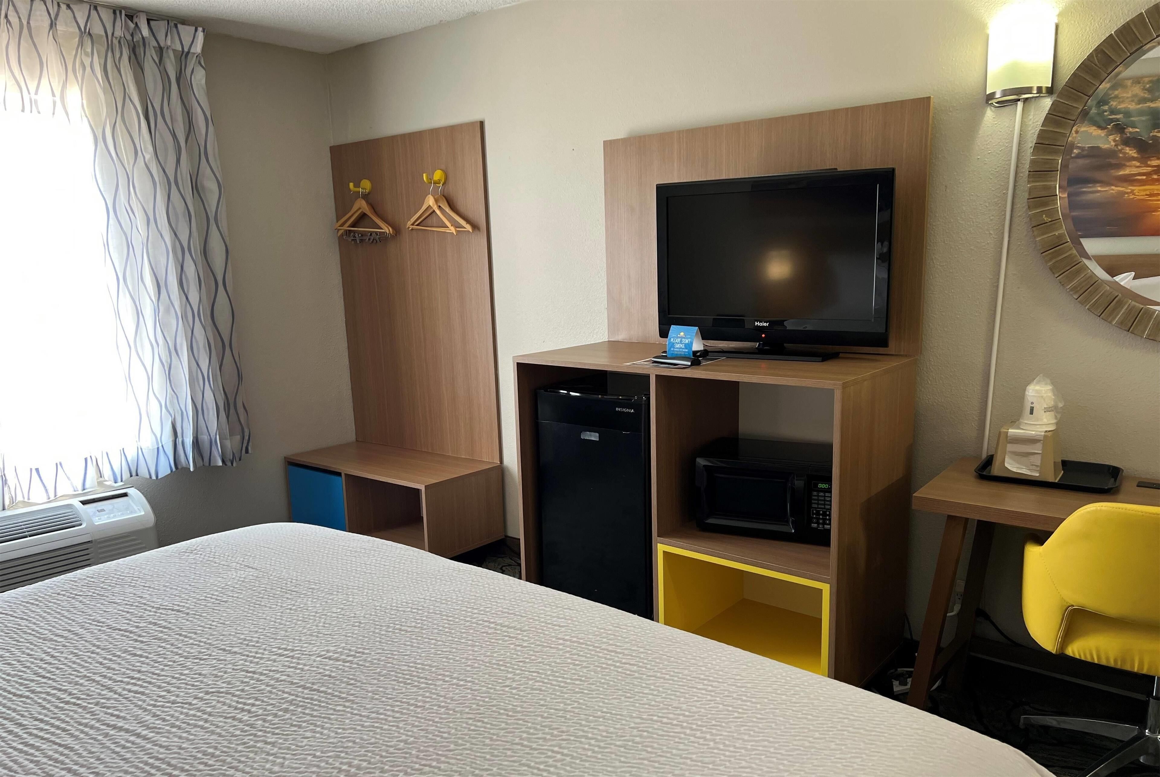 Quarto de hotel com cama, televisão, micro-ondas, mini-geladeira, escrivaninha com cadeira e janela com cortinas.