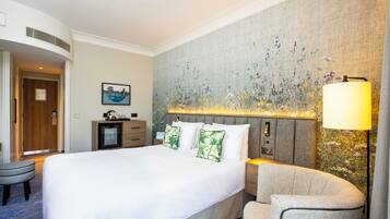 A hotel room with a large bed, a nightstand, a lamp, a chair, a desk, and a painting on the wall.