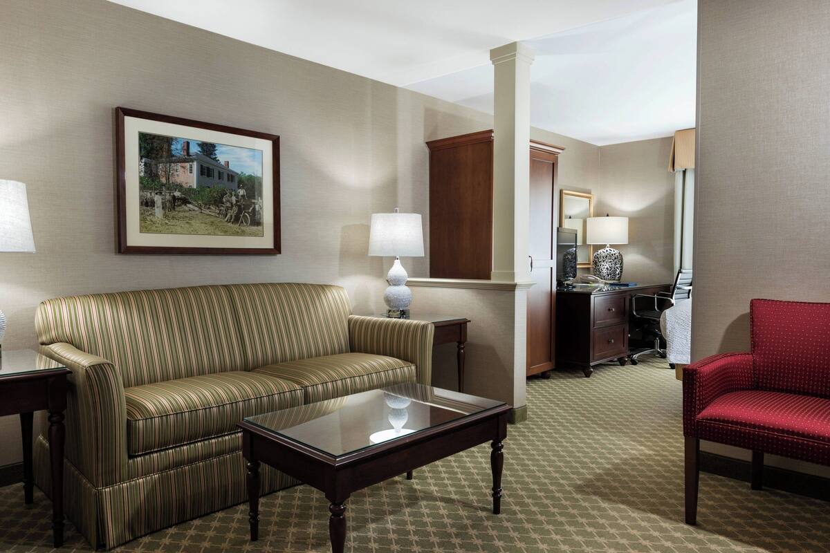 A hotel room with a striped sofa, a glass-top coffee table, a red chair, a desk with a lamp, and a framed picture on the wall.
