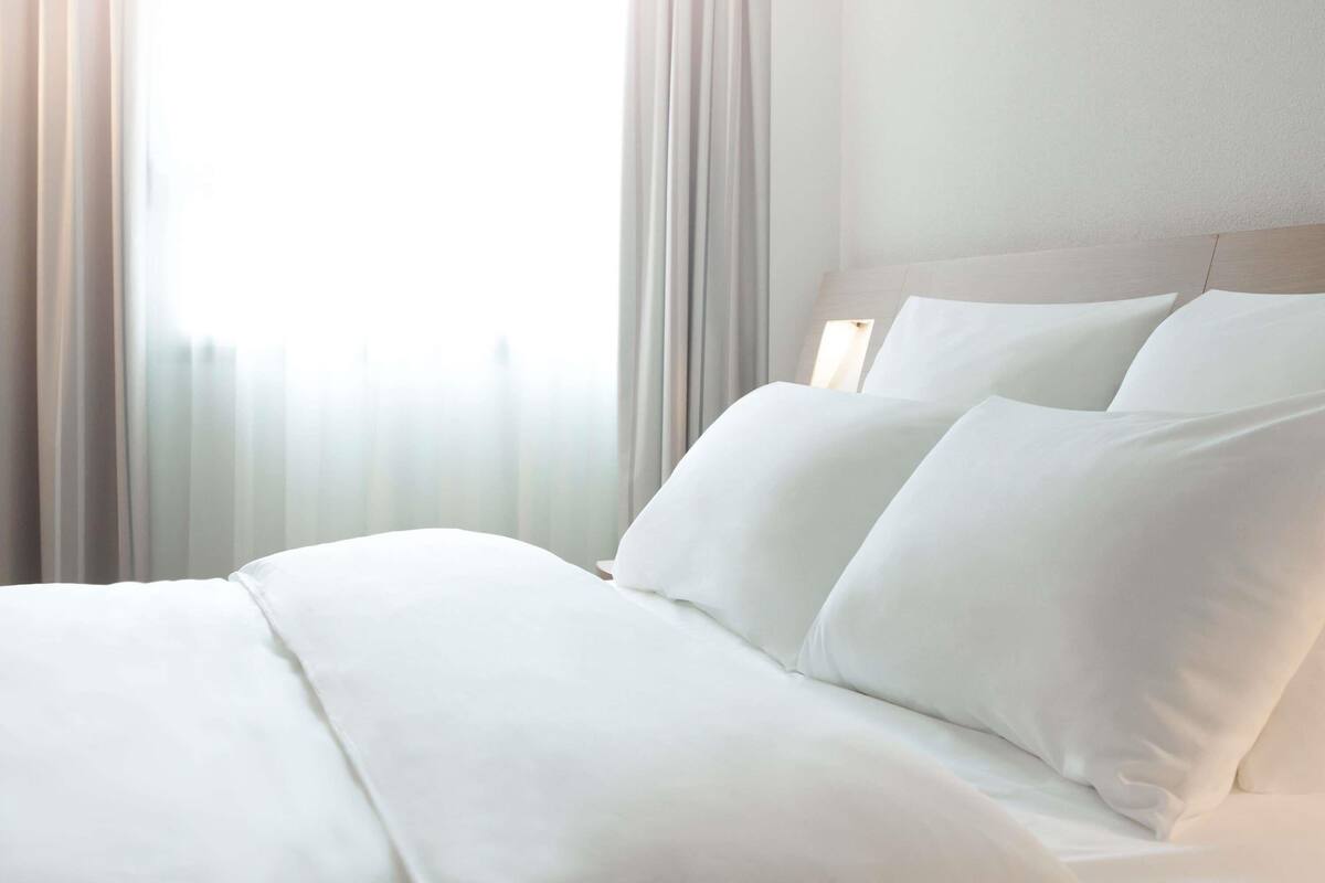A neatly made bed with white linens, a wooden headboard, and sheer curtains allowing natural light.