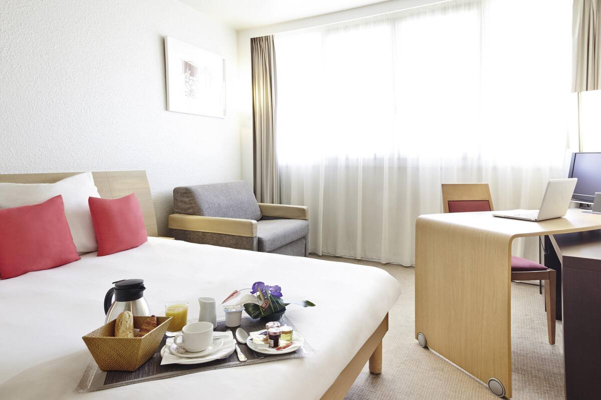 A hotel room with a bed, a desk with a laptop, a chair, and a breakfast tray with coffee, juice, and pastries.