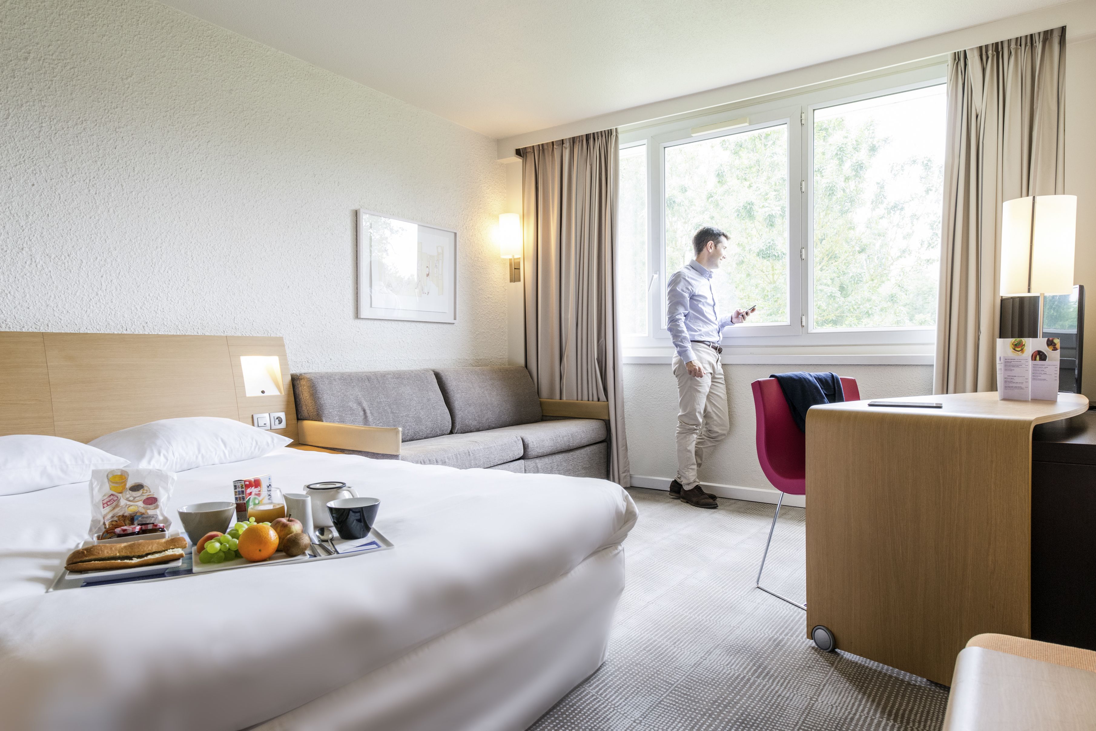 Et hotelværelse med seng, skrivebord, stol, sofa, vindue og et bakke med morgenmad.