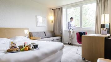 Et hotelværelse med seng, skrivebord, stol, sofa, vindue og et bakke med morgenmad.