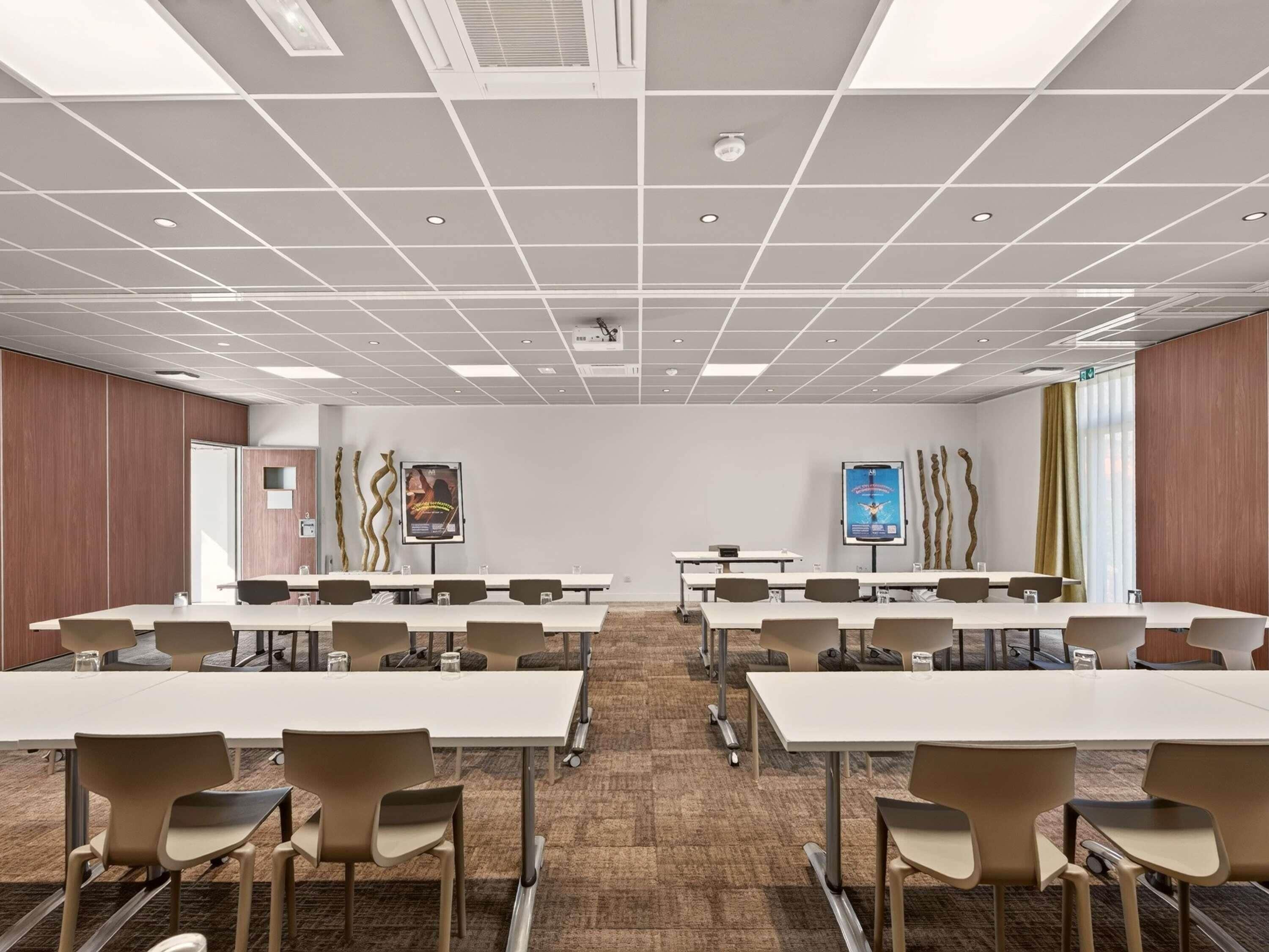 Un salón de conferencias con varias mesas y sillas, piso alfombrado y techo blanco con luces empotradas.