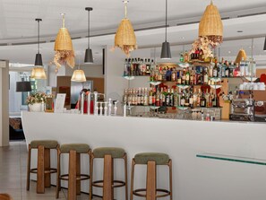 Zona de bar con mostrador, taburetes, lámparas de mimbre colgantes y estantes llenos de botellas.