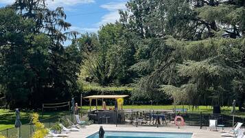 A swimming pool surrounded by a deck with lounge chairs, a covered seating area, and a fenced garden.