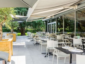 An outdoor dining area with tables and chairs, large windows, and a covered section with a canopy. - Novotel Pau Pyrénées (Lescar)