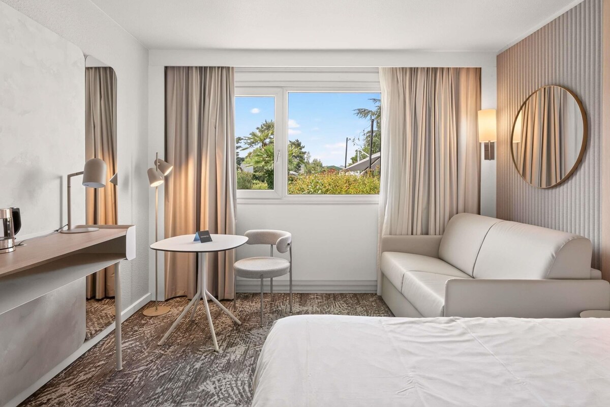 Habitación de hotel con cama, sofá, escritorio con silla, una mesita y ventana con cortinas.