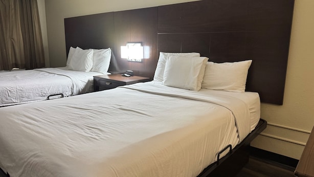 A hotel room with two beds, each with white linens and pillows, a wooden headboard, a nightstand with a lamp, and a window with curtains.