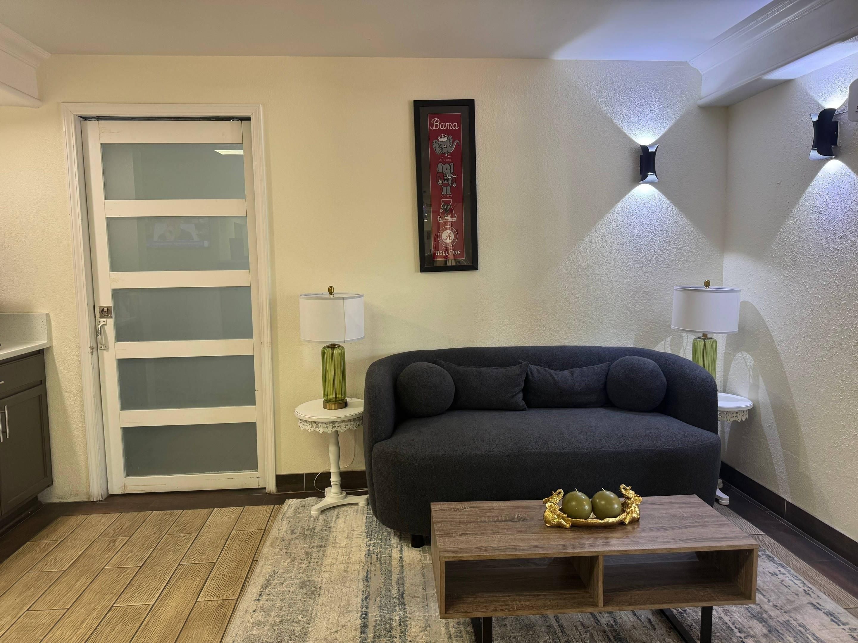 Ein Wohnzimmer mit einem dunklen Sofa, einem kleinen runden Tisch mit einem dekorativen Gegenstand, einer weißen Tür mit Milchglasfenstern und zwei Wandleuchten.