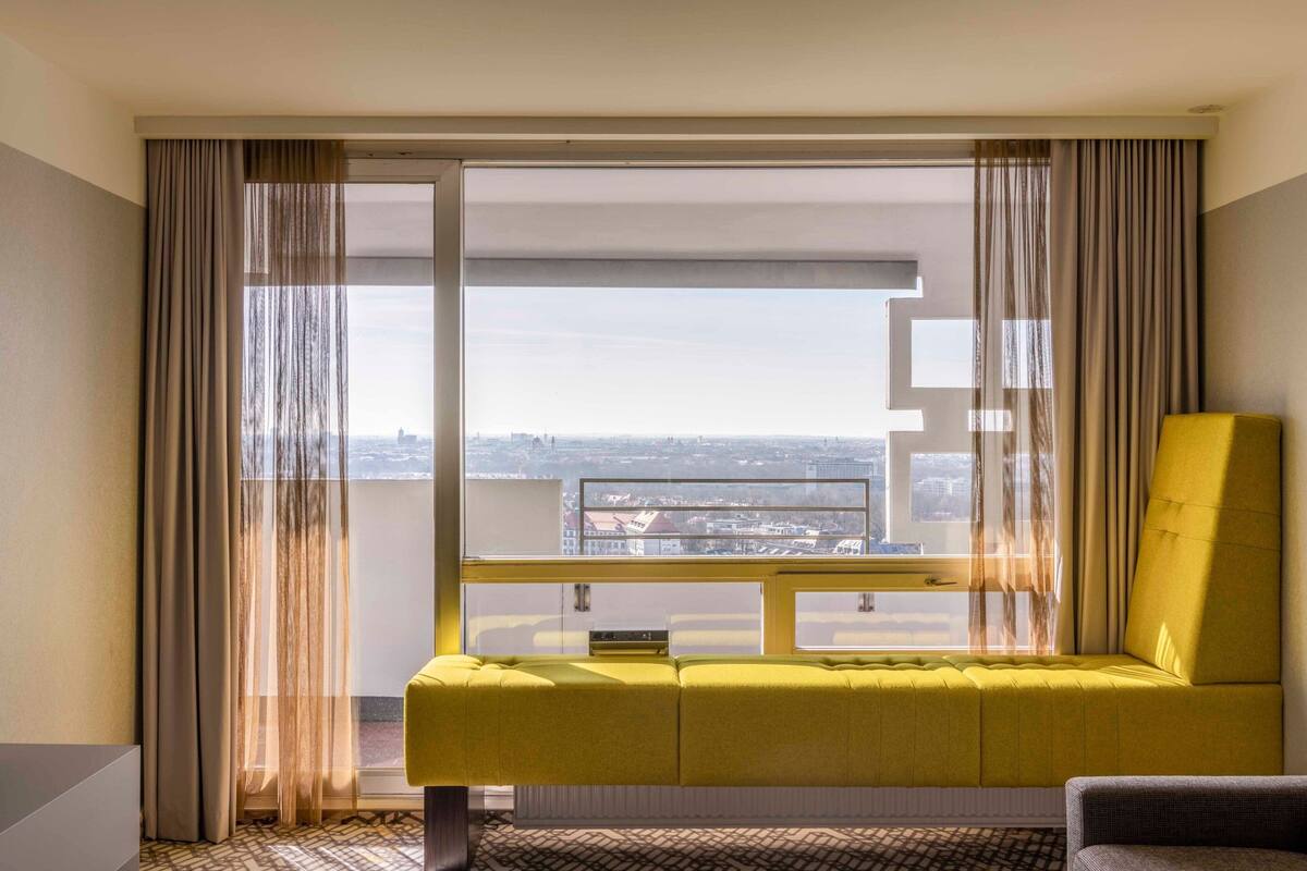 Ein modernes Hotelzimmer mit einem großen Fenster, einem gelben Sofa und Blick auf die Stadtlandschaft.