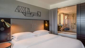 Ein modernes Hotelzimmer mit einem großen Bett, einem Nachttisch, einer Wandlampe und einem durch eine Glasabtrennung sichtbaren Badezimmer.