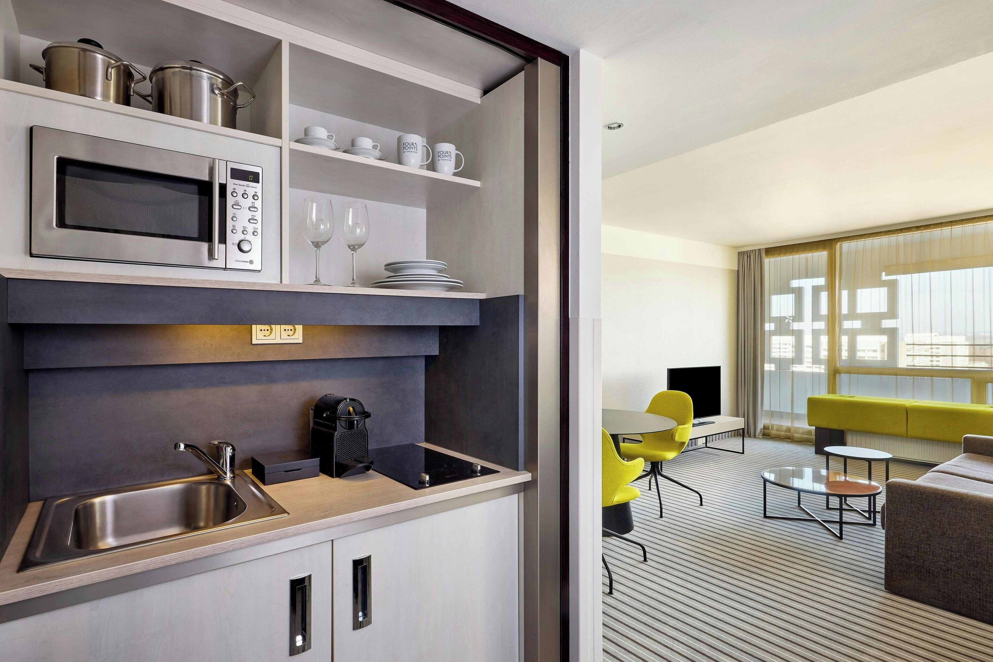 A modern kitchen with a built-in microwave, sink, and coffee maker. Adjacent to the kitchen is a living area with a sofa, coffee table, and yellow chairs.
